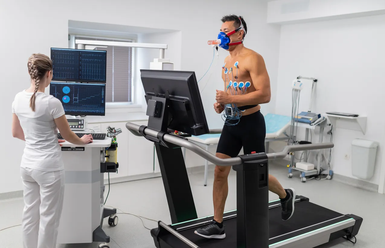A patient on a treadmill undergoing a traditional in-clinic CPET cardiopulmonary exercise stress test, wearing a gas-exchange mask and ECG electrodes while a clinician monitors the readouts